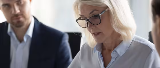 A focused woman with glasses, wearing a light blue blouse, is engaged in conversation during a business meeting. A man in a suit listens attentively.