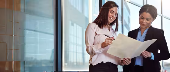 Two women in business attire reviewing a document in a modern office.