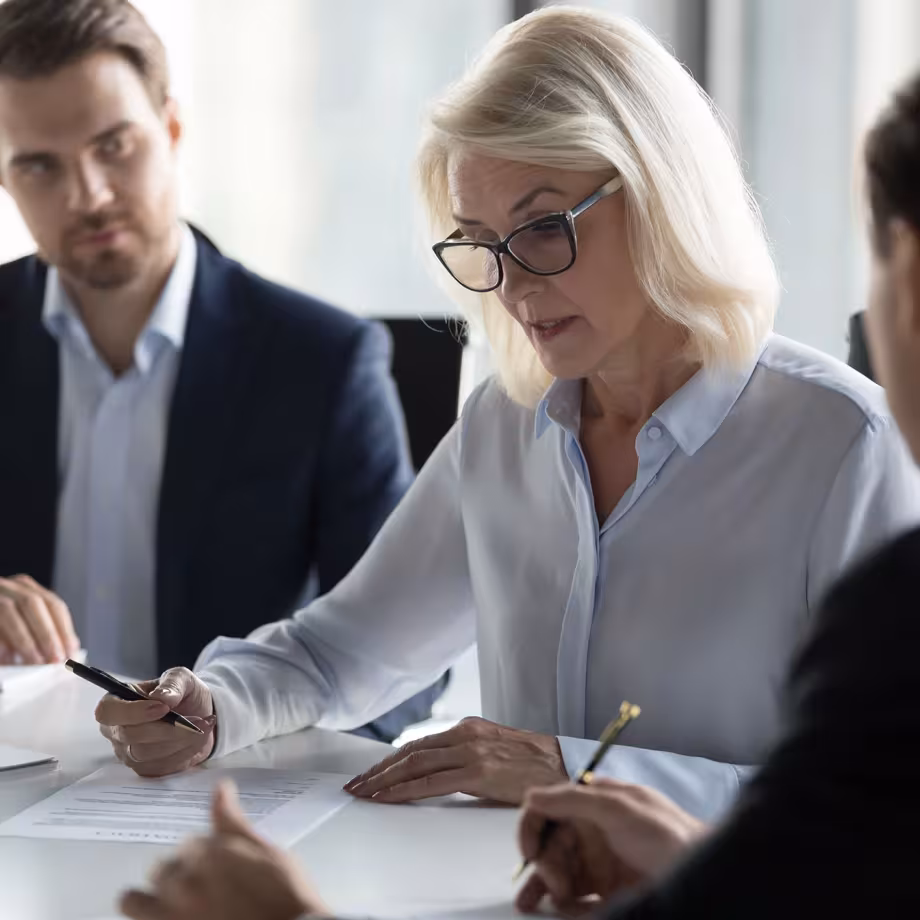 A woman with glasses signs papers at a business meeting, focused and serious. Two colleagues, a man and another partially visible person, watch.