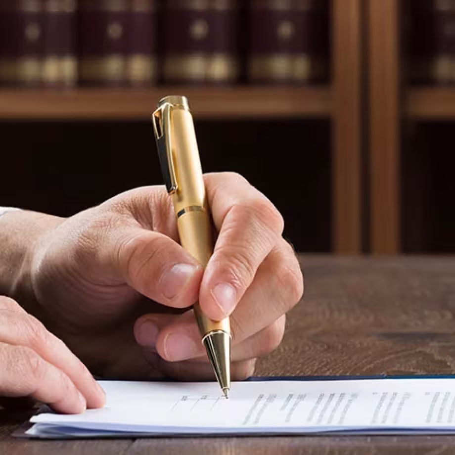 A hand with a gold pen signs a document on a wooden table, with law books in the background.