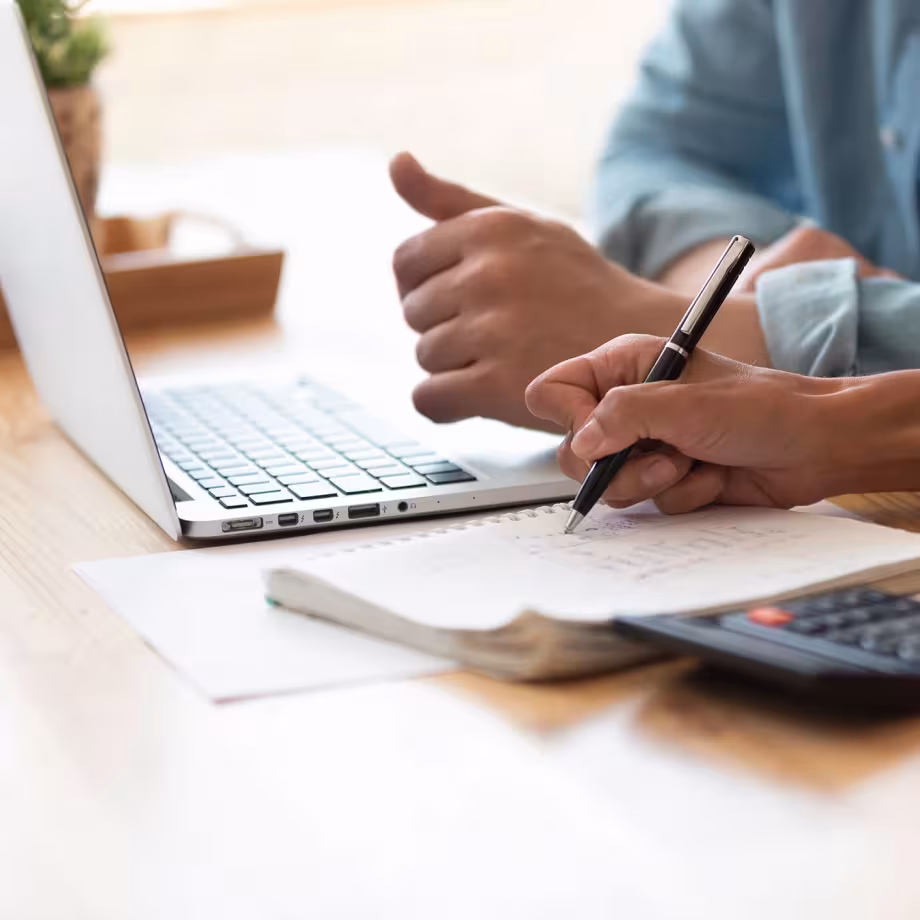Two people collaborate at a desk with a laptop, taking notes and using a calculator.