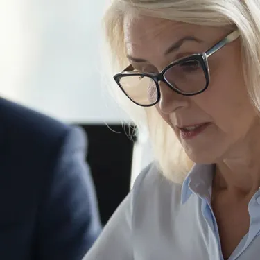 A focused woman with glasses, wearing a light blue blouse, is engaged in conversation during a business meeting. A man in a suit listens attentively.