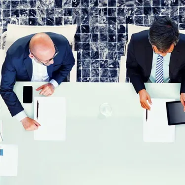 Four people sit around a glass conference table with papers, notebooks, coffee cups, and tablets, engaged in a business meeting, viewed from above.