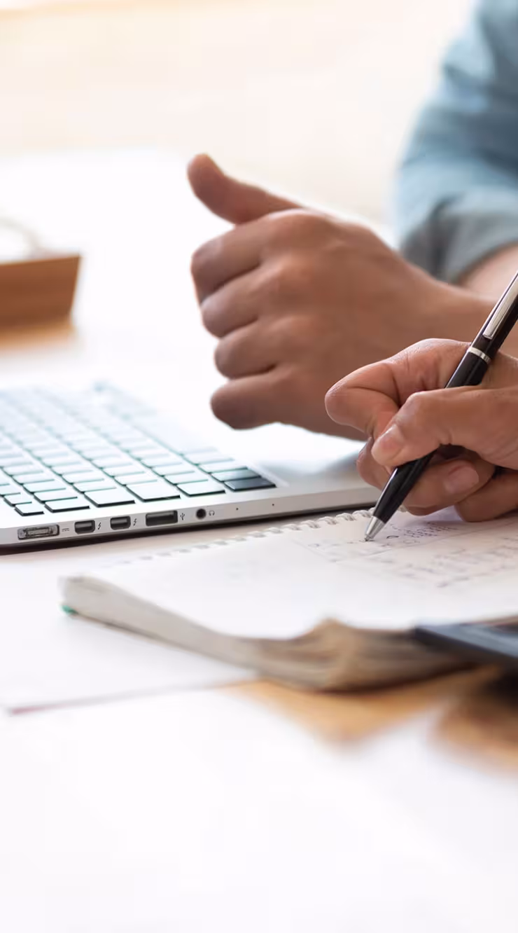Two people collaborate at a desk with a laptop, taking notes and using a calculator.